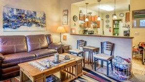 Una sala de estar con un sofá de cuero marrón, una mesa de centro de madera y un comedor con sillas y una pequeña mesa. Un espejo grande refleja la zona de la cocina con armarios y un refrigerador.