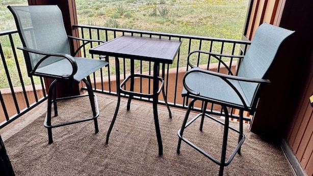 Een overdekt balkon met twee stoelen en een tafel, met uitzicht op een grasveld en gebouwen in de verte.