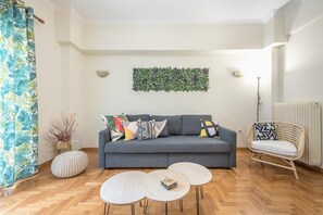 Ein Wohnzimmer mit grauem Sofa, zwei runden Tischen, einem Korbstuhl, einer grünen Wanddekoration aus Blättern und einem gemusterten Vorhang.