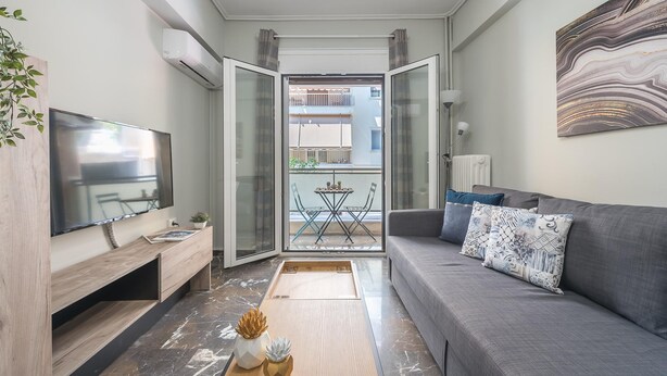 Una sala moderna con un sofá gris, un televisor de pantalla plana, una mesa de centro de madera y vistas a un balcón exterior con mesa y sillas.