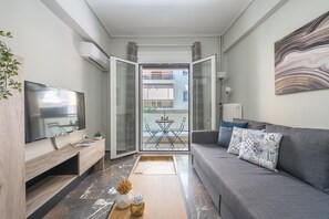Una sala moderna con un sofá gris, un televisor de pantalla plana, una mesa de centro de madera y vistas a un balcón exterior con mesa y sillas.