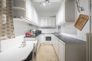 Une cuisine compacte avec des meubles blancs, un plan de travail noir, une bouilloire rouge et une planche à découper en bois portant une inscription.
