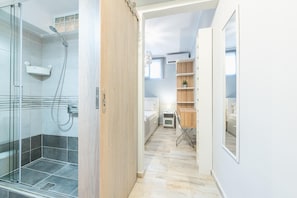 A modern bathroom with a glass shower enclosure, a wooden vanity, and a small bedside table with a lamp.
