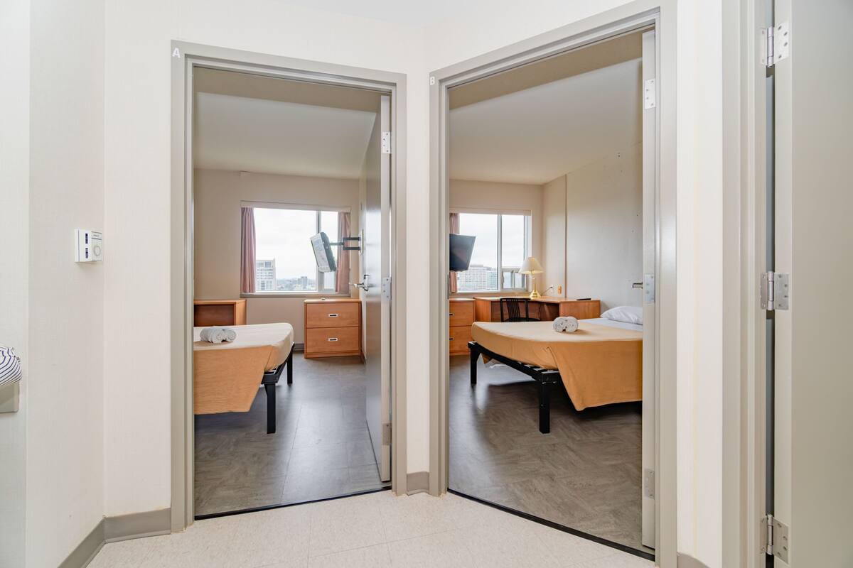 A hotel room with a bed, desk, and chair, a window with a view of buildings, and a door leading to another room.