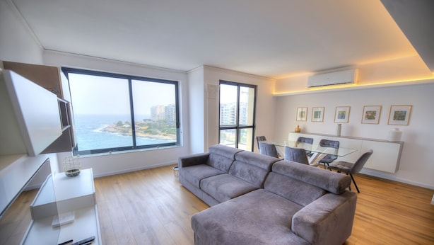 A modern living room with a large sectional sofa, a glass dining table, and a view of the ocean through large windows. - Bright & cozy 3bedrooms with stunning views DLAP1-1 (Sliema)
