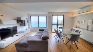 A modern living room with a large flat-screen TV, a grey sectional sofa, a glass dining table with chairs, and a view of the sea through large windows. - Bright & cozy 3bedrooms with stunning views DLAP1-1 (Sliema)