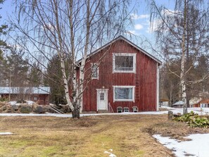 Ein rotes Bauernhaus mit weißer Tür und Fenstern, umgeben von Bäumen und einem grasbewachsenen Feld mit Schneeflecken.