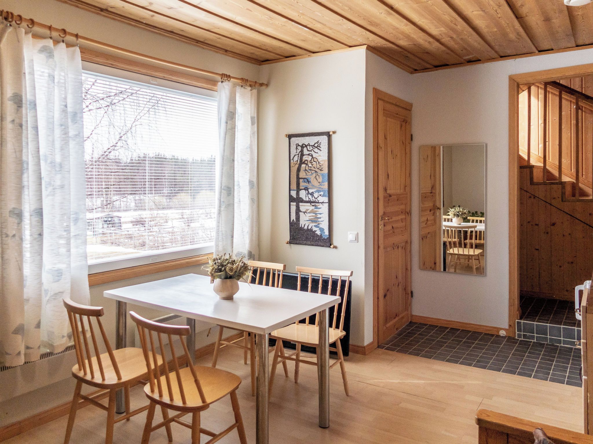Un área de comedor con una mesa y sillas, una ventana con cortinas, un cuadro en la pared y una escalera que conduce a otra habitación.