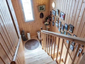Un escalier en bois, doté d’une rampe, mène à un palier agrémenté d’un tapis, d’un vase de fleurs et d’un mur orné de photographies encadrées.