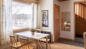 Ruokailutila, jossa on valkoinen pöytä ja puiset tuolit, ikkuna, jossa on verhot, seinällä oleva taulu ja puinen portaikko, joka johtaa toiseen huoneeseen.
