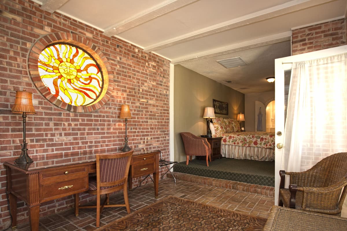 A room with a brick wall, a large circular window with a sun design, a desk with two chairs, a bed with a floral bedspread, and a door with a curtain.