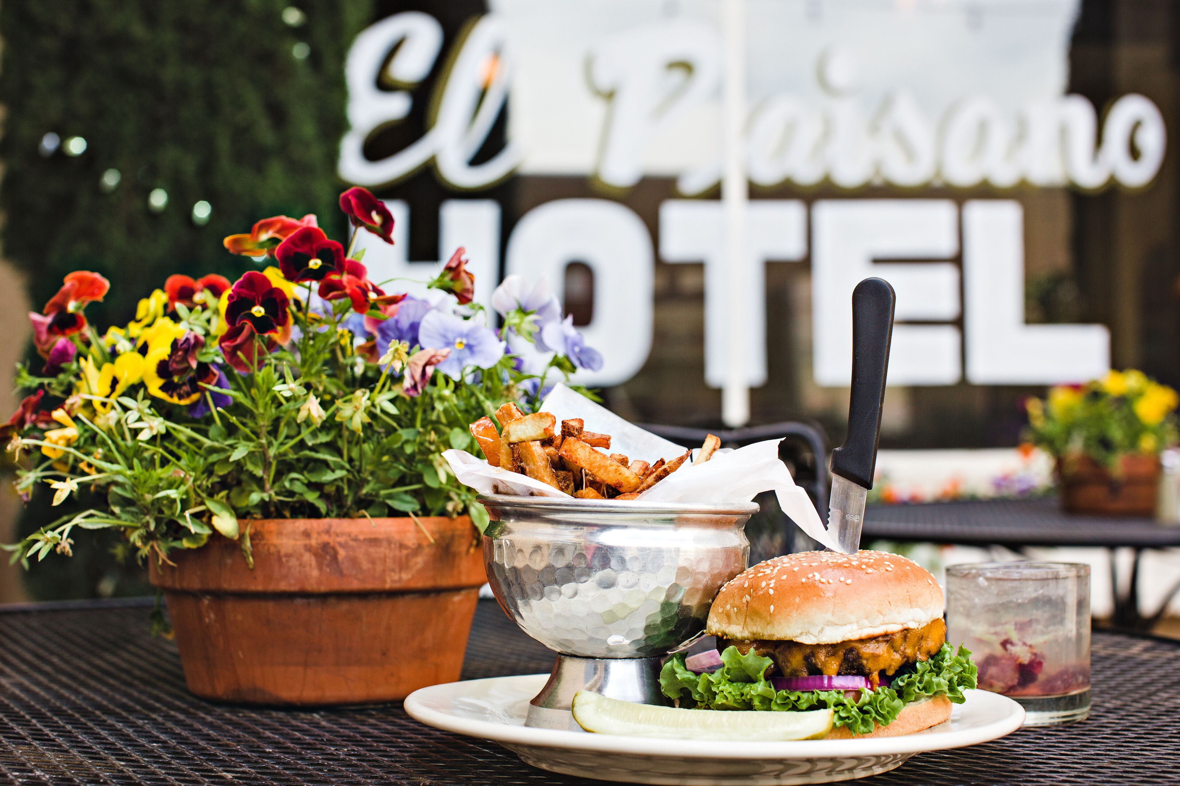 A dining table with a burger, fries, and a drink, with a sign reading "El Paisano Hotel" in the background.