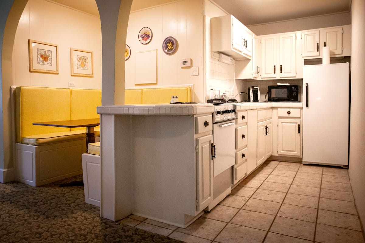 A kitchen with white cabinets, a microwave, an oven, and a refrigerator. There are framed pictures on the wall and a yellow bench in the corner.
