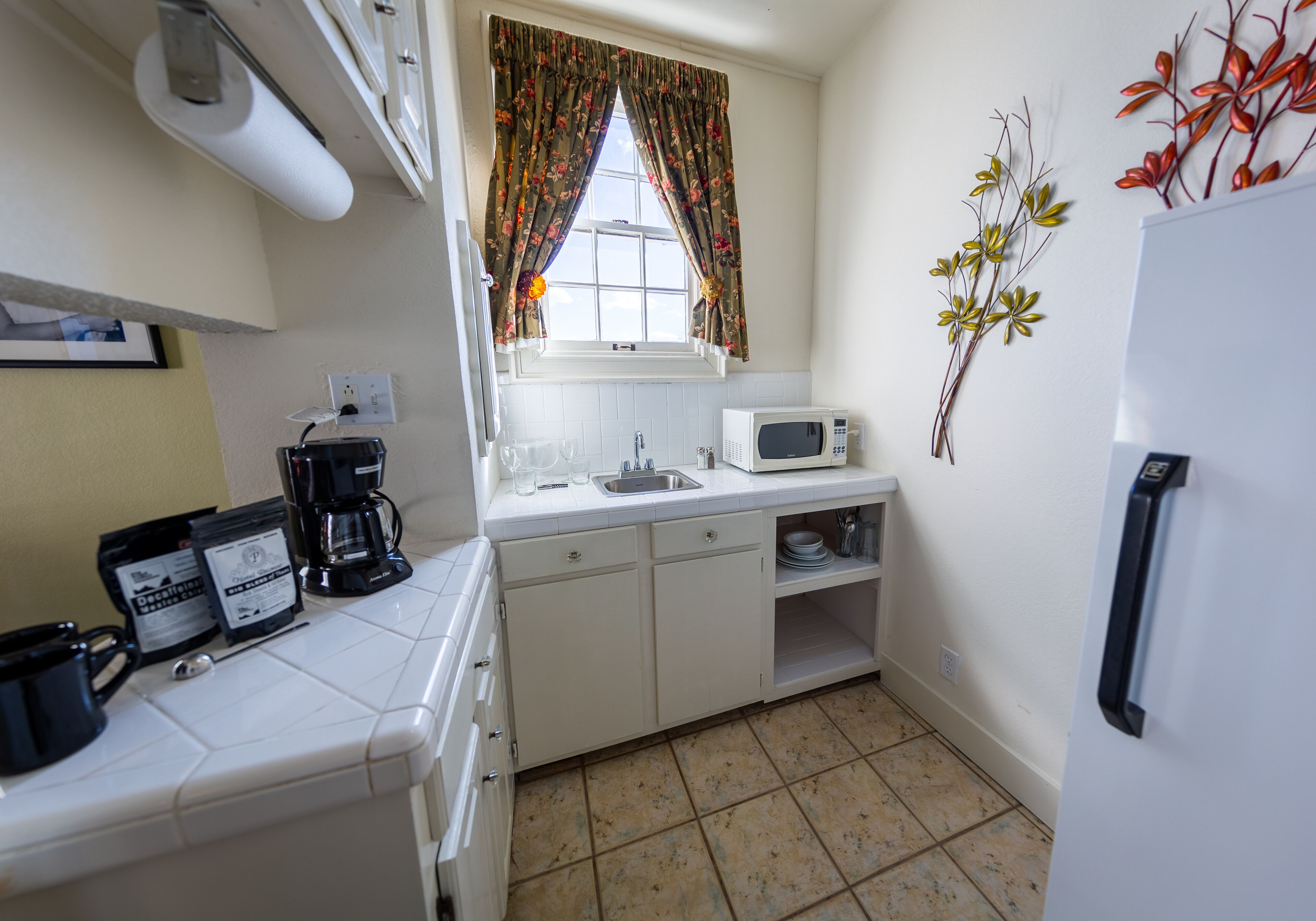 A compact kitchen with white cabinets, a microwave, a coffee maker, and a window with floral curtains.