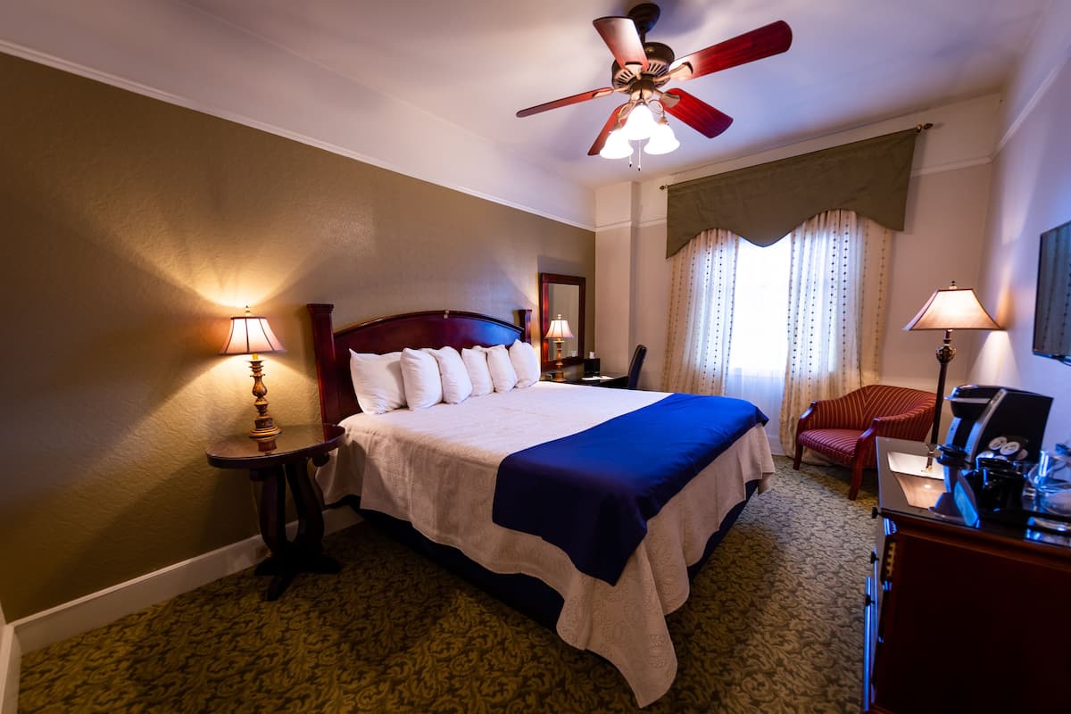 A hotel room with a large bed, a ceiling fan, a desk with a coffee machine, and a chair.
