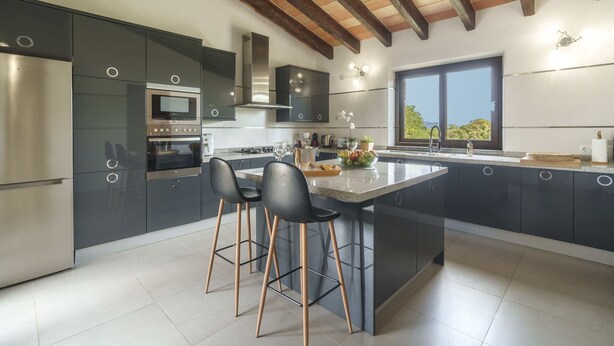 A modern kitchen with dark cabinets, a large island with bar stools, a built-in oven, and a window providing a view of the outdoors. - Bellpuig by Interhome (Artà)