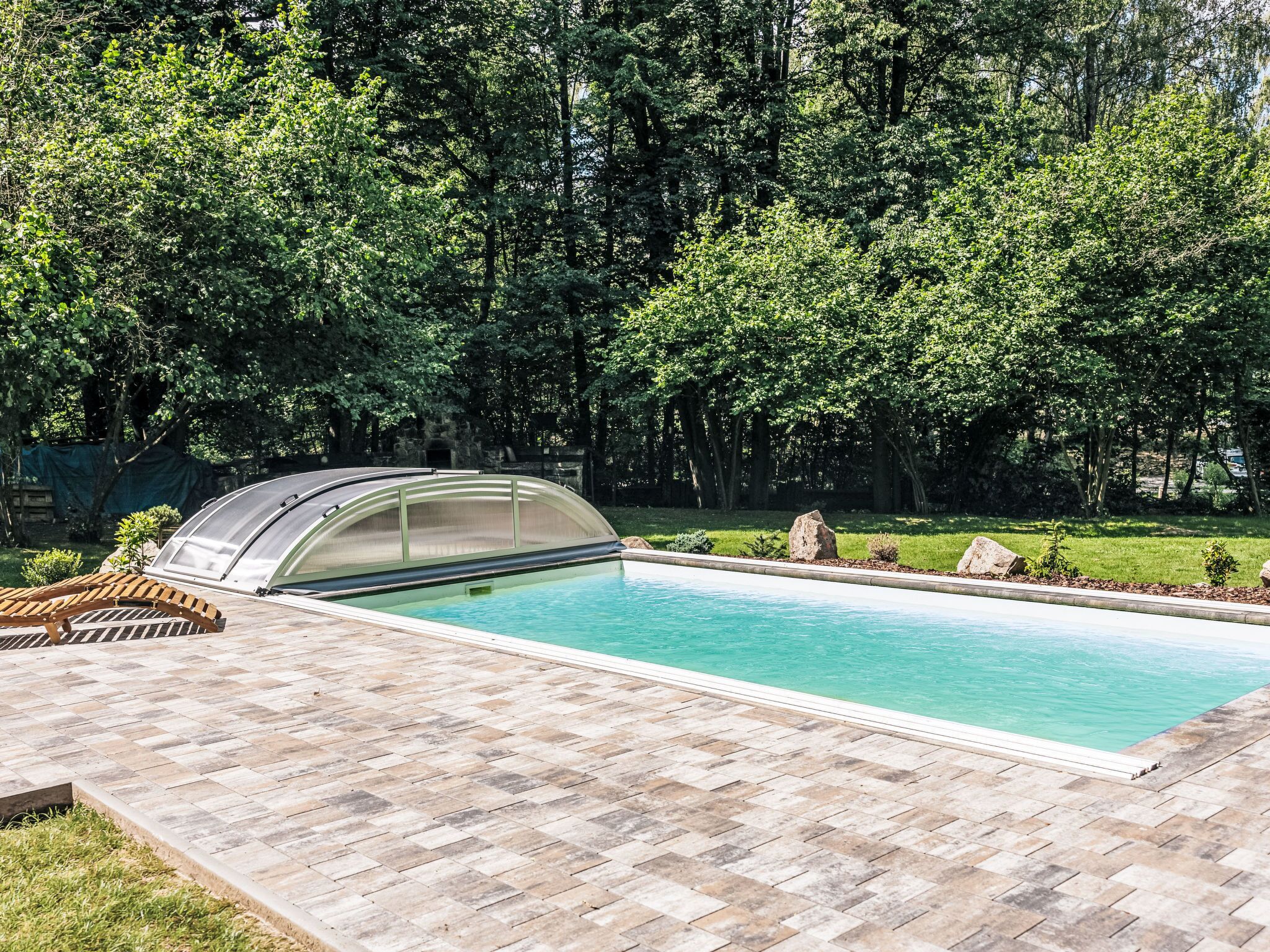 A swimming pool with a covered area, surrounded by a paved deck and lush greenery.