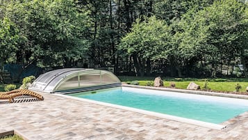 A swimming pool with a covered area, surrounded by a paved deck and lush greenery.