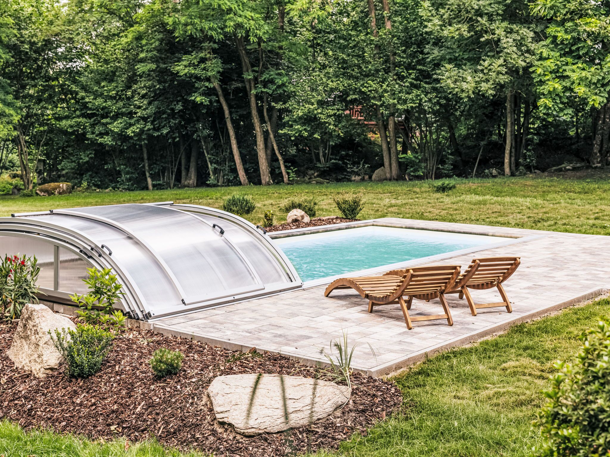 Une piscine extérieure couverte, dotée d’un espace patio, de chaises longues en bois et entourée d’un écrin de verdure.