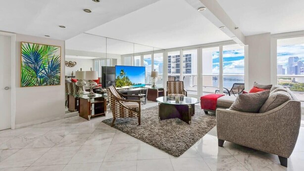 A spacious living room with a large TV, comfortable seating, and a city view through large windows. - Classic Waterfront Apt | Bay Views | StayCozy (Miami)