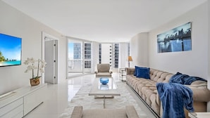 A modern living room with a sofa, coffee table, and a TV. There is a large window with a view of buildings. - Bay Overlooking Balcony | 400 Mbps WiFi | StayCozy (Miami)