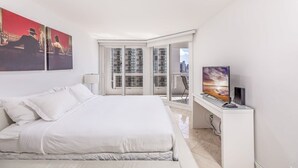 A modern bedroom with a large bed, a desk with a computer, and a city view through floor-to-ceiling windows. - Bayfront Condo | Elevated amenities | StayCozy (Miami)