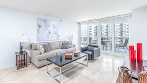 A modern living room with a sofa, coffee table, and chairs, a large window with a city view, and a painting on the wall. - Bayfront Condo | Elevated amenities | StayCozy (Miami)