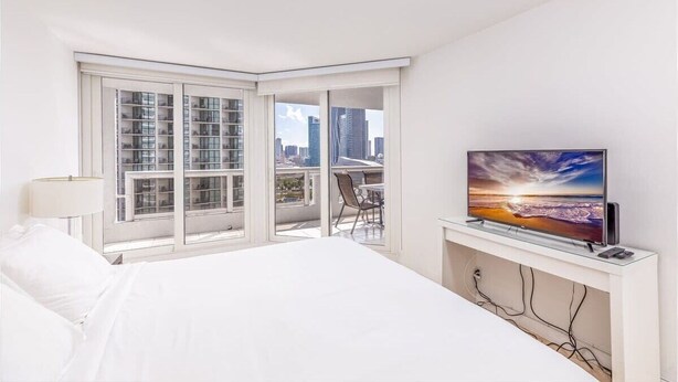 A hotel room with a large bed, a TV on a stand, a balcony with a city view, and a chair. - Bayfront Condo | Elevated amenities | StayCozy (Miami)