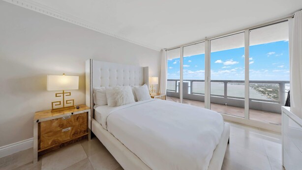 A bedroom with a large bed, a nightstand, a lamp, and a view of the sea through a balcony. - Private Balcony | Biscayne Bay View | StayCozy (Miami)