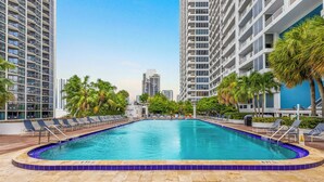 A swimming pool with lounge chairs, surrounded by tall buildings and palm trees. - Private Balcony | Biscayne Bay View | StayCozy (Miami)