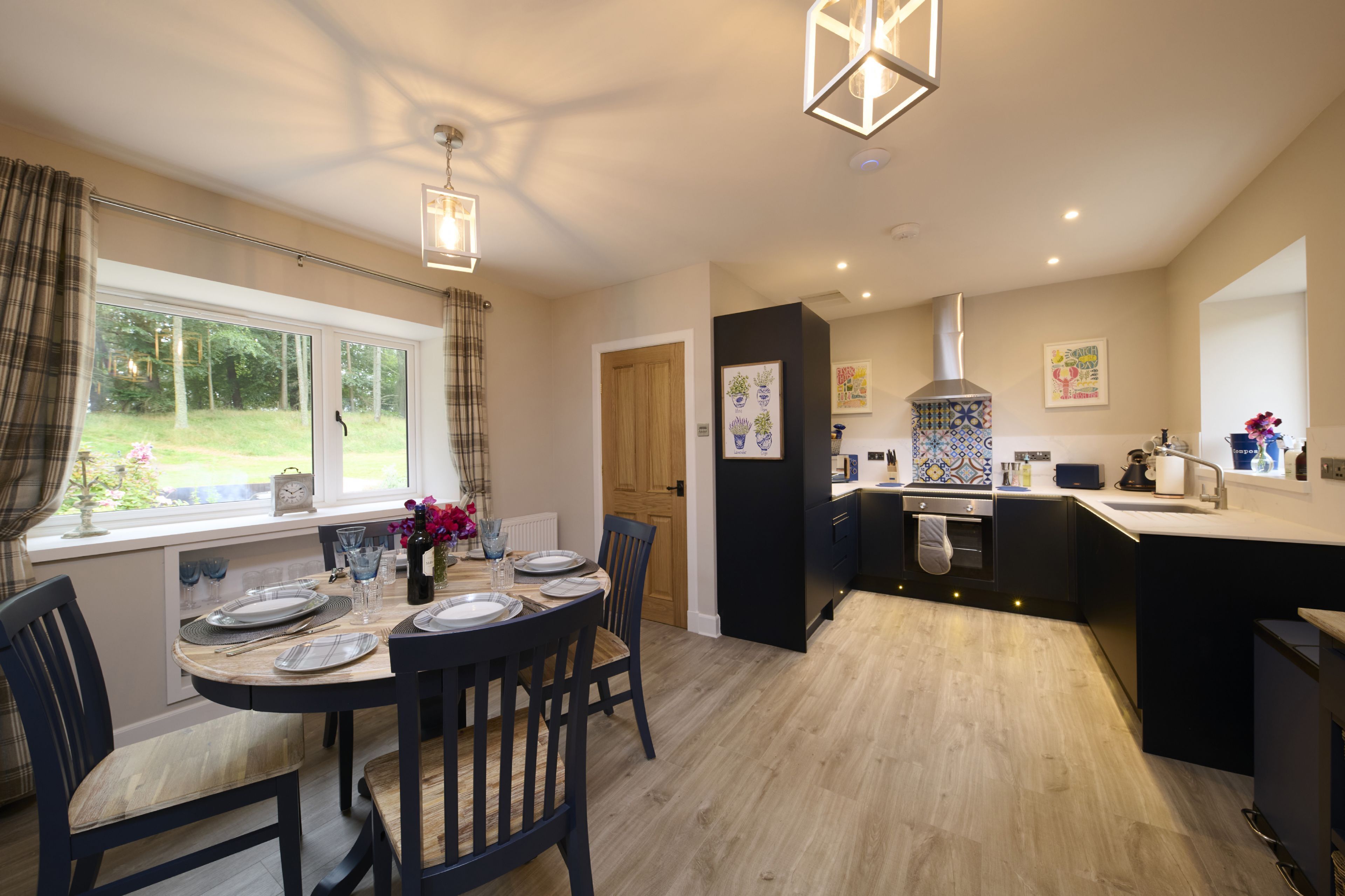 A modern kitchen with a central island, a dining area with a round table, and a view of the garden through large windows.