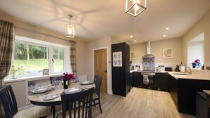 A modern kitchen with a central island, a dining area with a round table, and a view of the garden through large windows. - Luxury Highland Hideaway with Hot Tub by LochNess (Inverness)