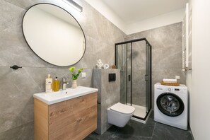 Features a sleek bathroom with a large mirror, washing machine, glass-enclosed shower, and a toilet.
