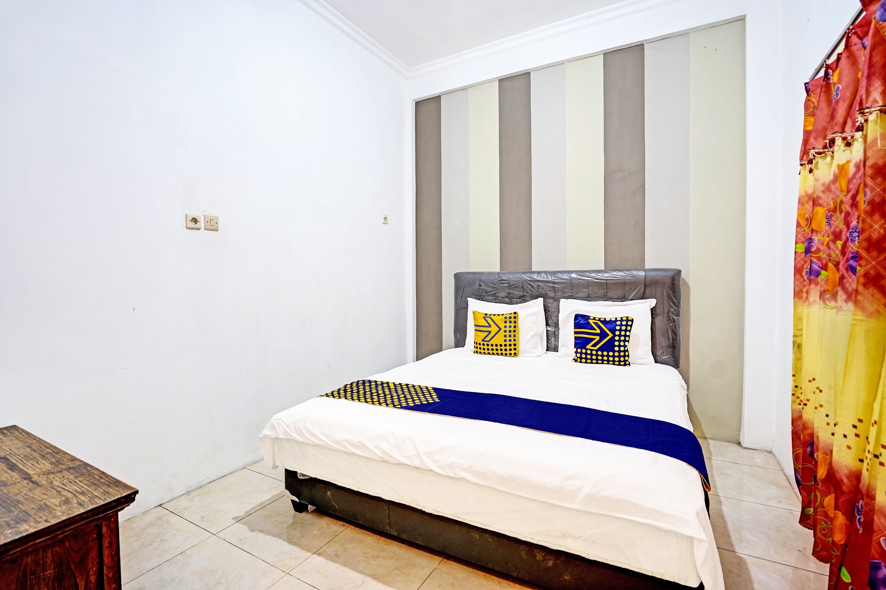 A bedroom with a bed, a wooden nightstand, a striped headboard, and a colorful curtain.