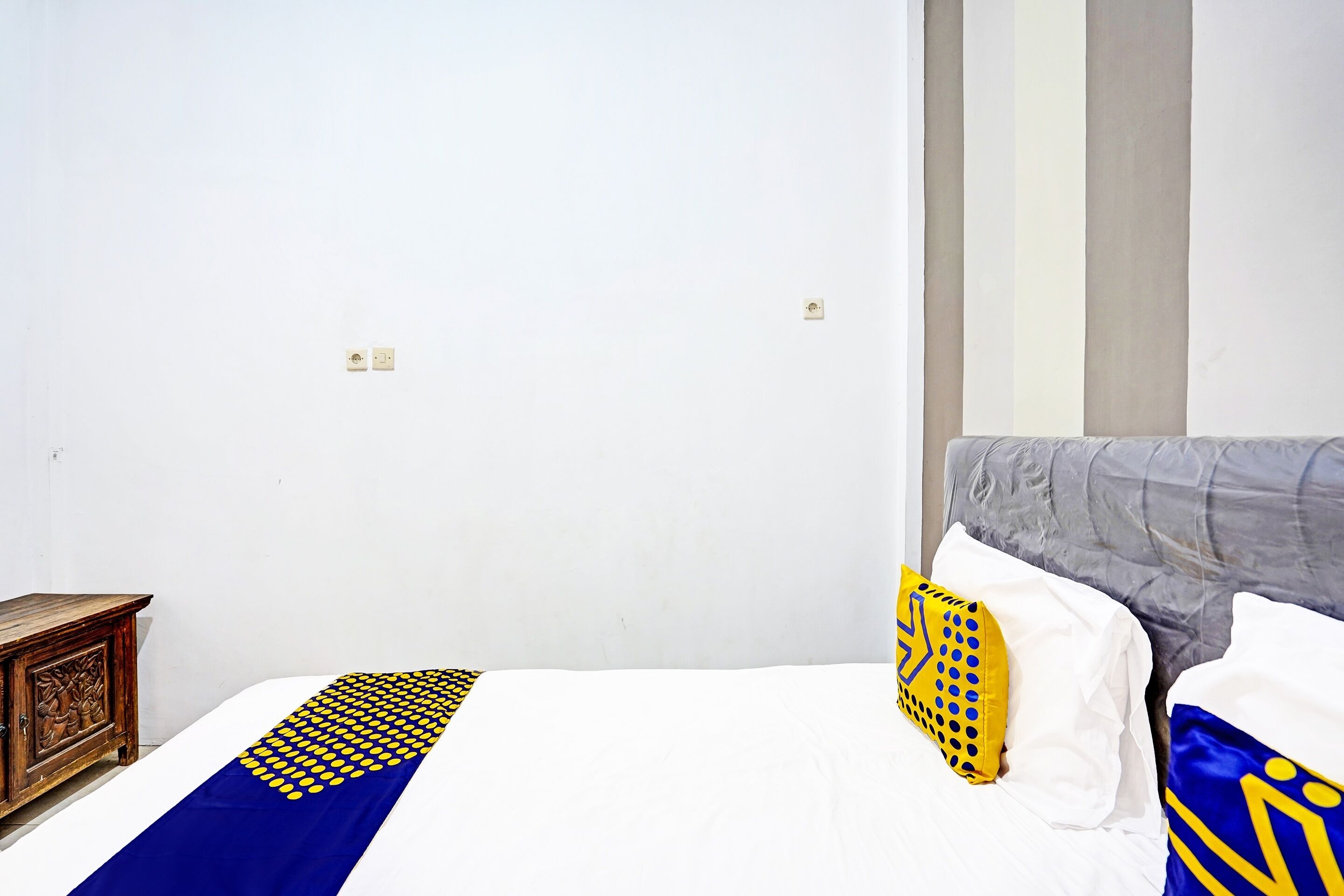 A hotel room with a bed, a wooden nightstand, and a plain white wall.