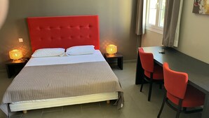 A hotel room with a red upholstered bed, two bedside lamps, a desk with a chair, and a window with curtains.