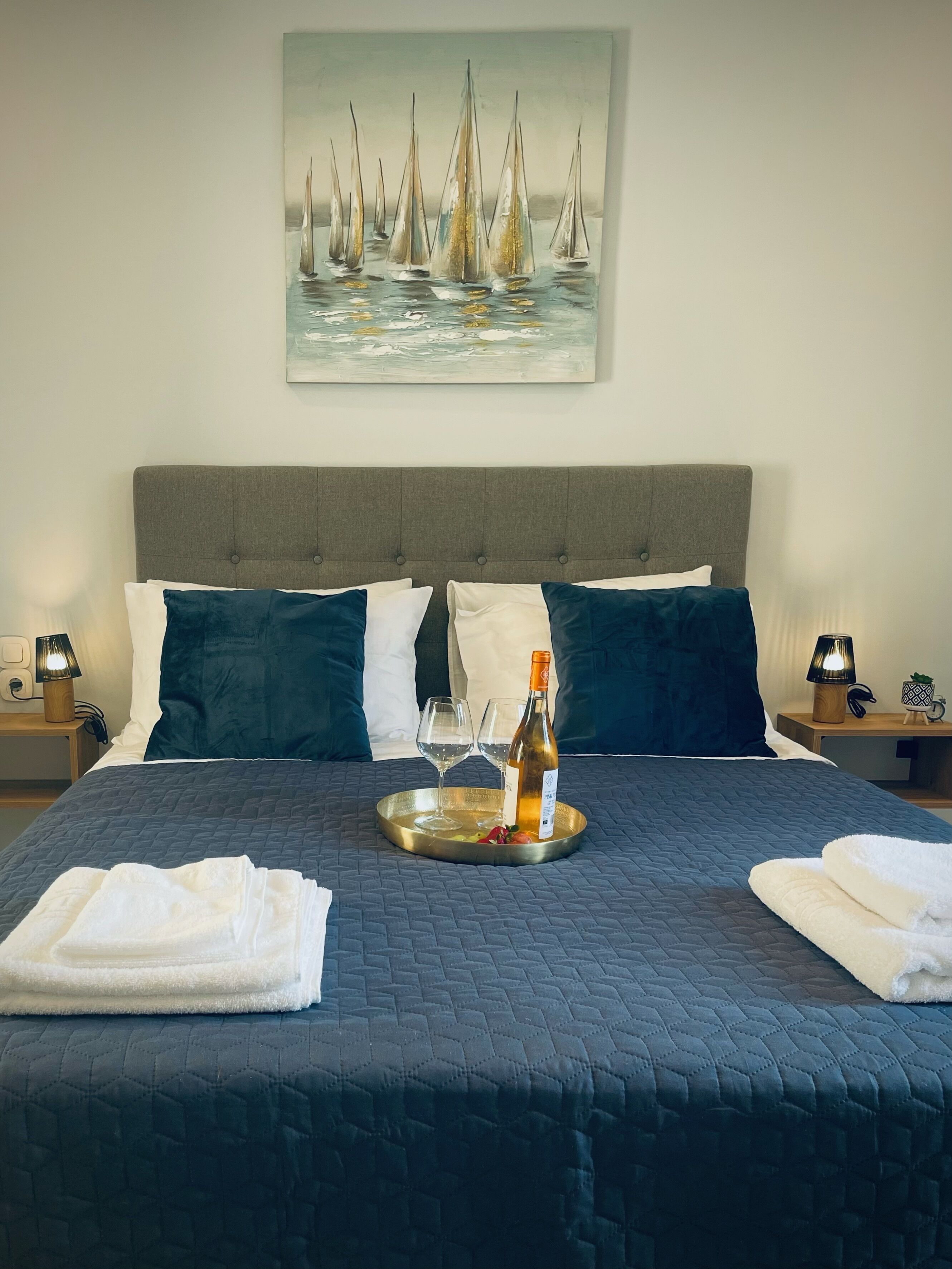 A neatly made bed with a dark blue quilt, two pillows, a tray with a bottle and glasses, and a painting of sailboats on the wall.