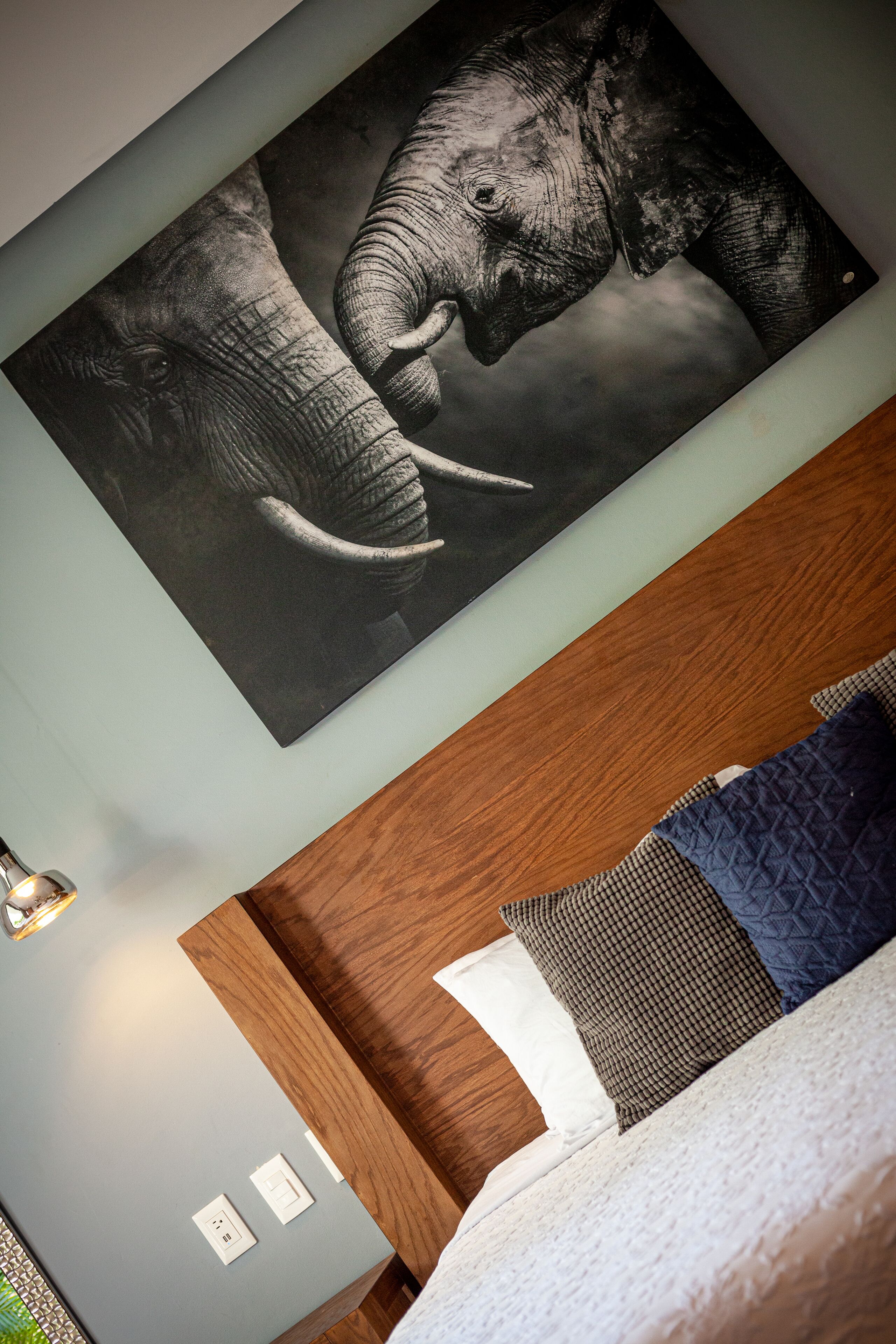 Quarto com cama, almofadas, uma obra de arte na parede com elefantes e cabeceira de madeira.
