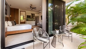Une chambre moderne avec un grand lit, un ventilateur de plafond et une porte-fenêtre coulissante donnant sur un balcon équipé de deux chaises et d’une table.