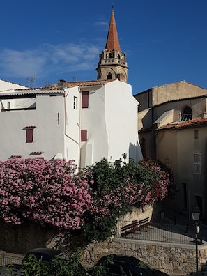Um edifício branco com telhado de telhas vermelhas, uma torre de sino e arbustos floridos em tons de rosa na frente.