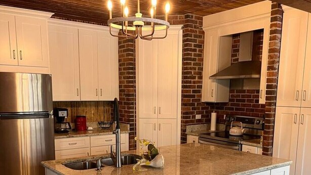 A modern kitchen with white cabinetry, a central island with a sink, a stainless steel refrigerator, and a brick accent wall.