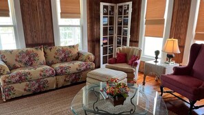 A living room with a floral sofa, a glass coffee table with a vase, a red armchair, and a wooden cabinet with glass doors.