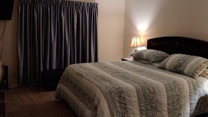 Een slaapkamer met een bed, nachtkastje, lamp, plafondventilator en een raam met gordijnen.