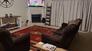 Een woonkamer met twee bruine fauteuils, een houten salontafel met tijdschriften, een open haard, een televisie waarop de Amerikaanse vlag wordt getoond, en een kamerbreed tapijt met een patroon.