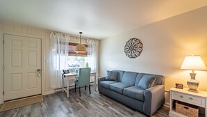 Ein Zimmer mit grauem Sofa, einem Schreibtisch mit Stuhl, einer Lampe und einer Wanddekoration.