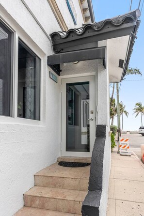 Un edificio blanco con un toldo negro sobre la entrada, una puerta de cristal y unos pocos escalones que conducen a ella.