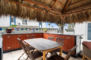 Un espacio al aire libre techado con mesa y sillas de madera, techo de paja y vista a un edificio con tejas de colores.