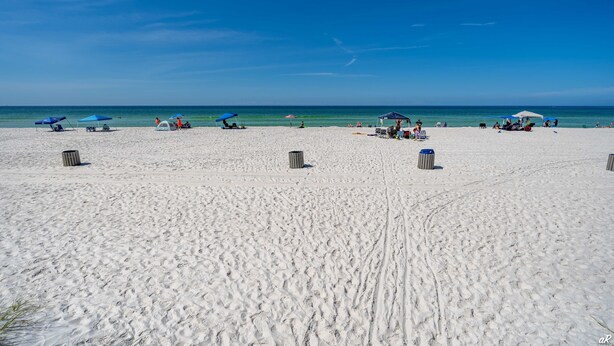 Ein weiter Sandstrand mit mehreren Sonnenschirmen und Liegestühlen, einigen Mülleimern und einem wolkenlosen, blauen Himmel.