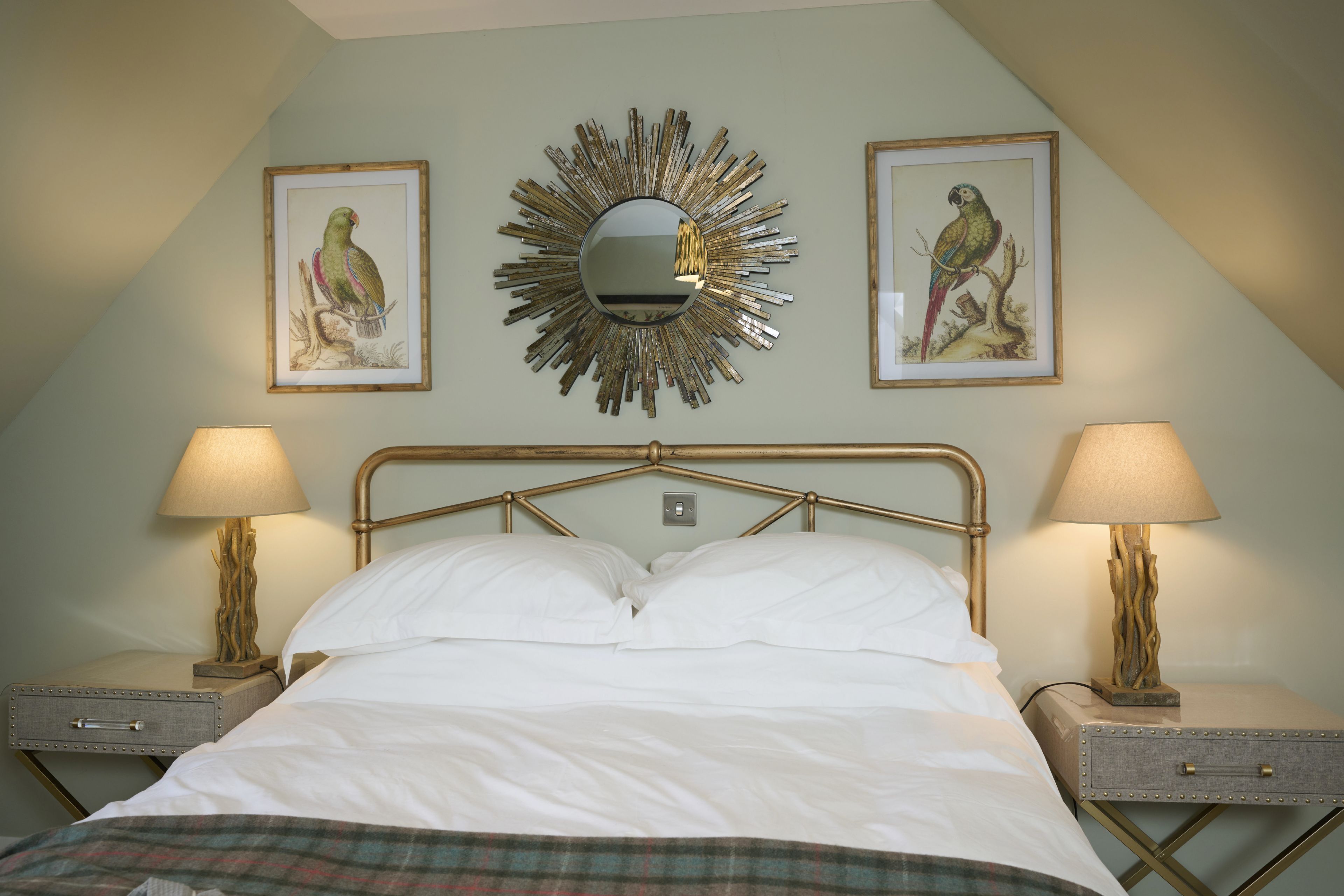 Une chambre à coucher avec un lit, deux tables de chevet avec des lampes, un miroir fixé au mur et deux tableaux encadrés représentant des perroquets.