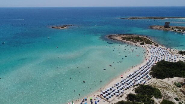 Vista aerea di una spiaggia con acque cristalline turchesi, una lunga distesa di ombrelloni e alcune imbarcazioni in lontananza.